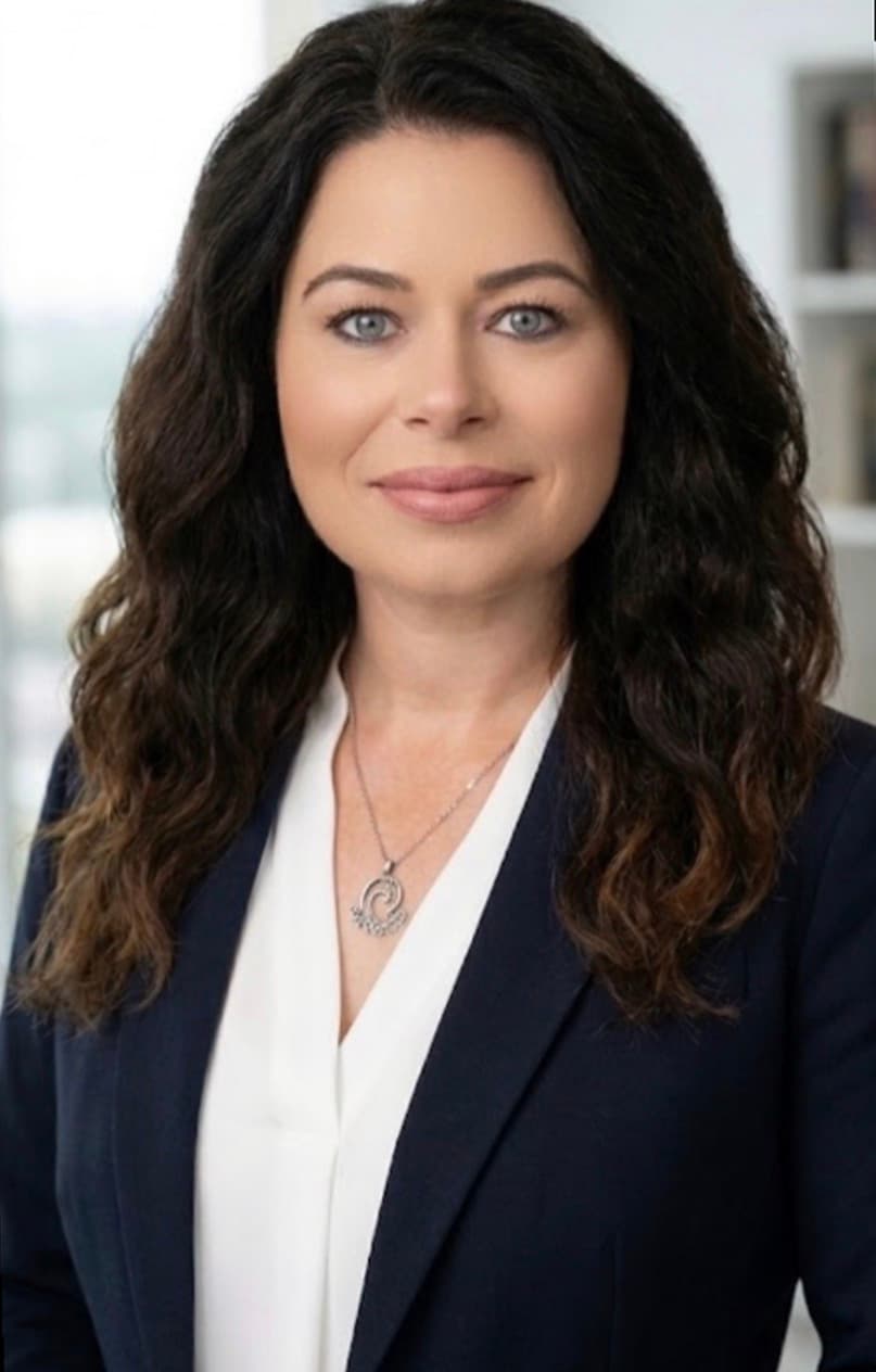 Woman with long dark wavy hair and blue eyes wearing a navy blazer and blouse.
